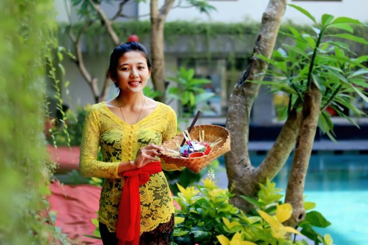 Pilihan 1 (Paling Direkomendasikan - Fokus SEO & Konteks): Seorang staf hotel di Bali menunjukkan skill wajib perhotelan yaitu keramahan dan 'service heart' sambil membawa canang sari.